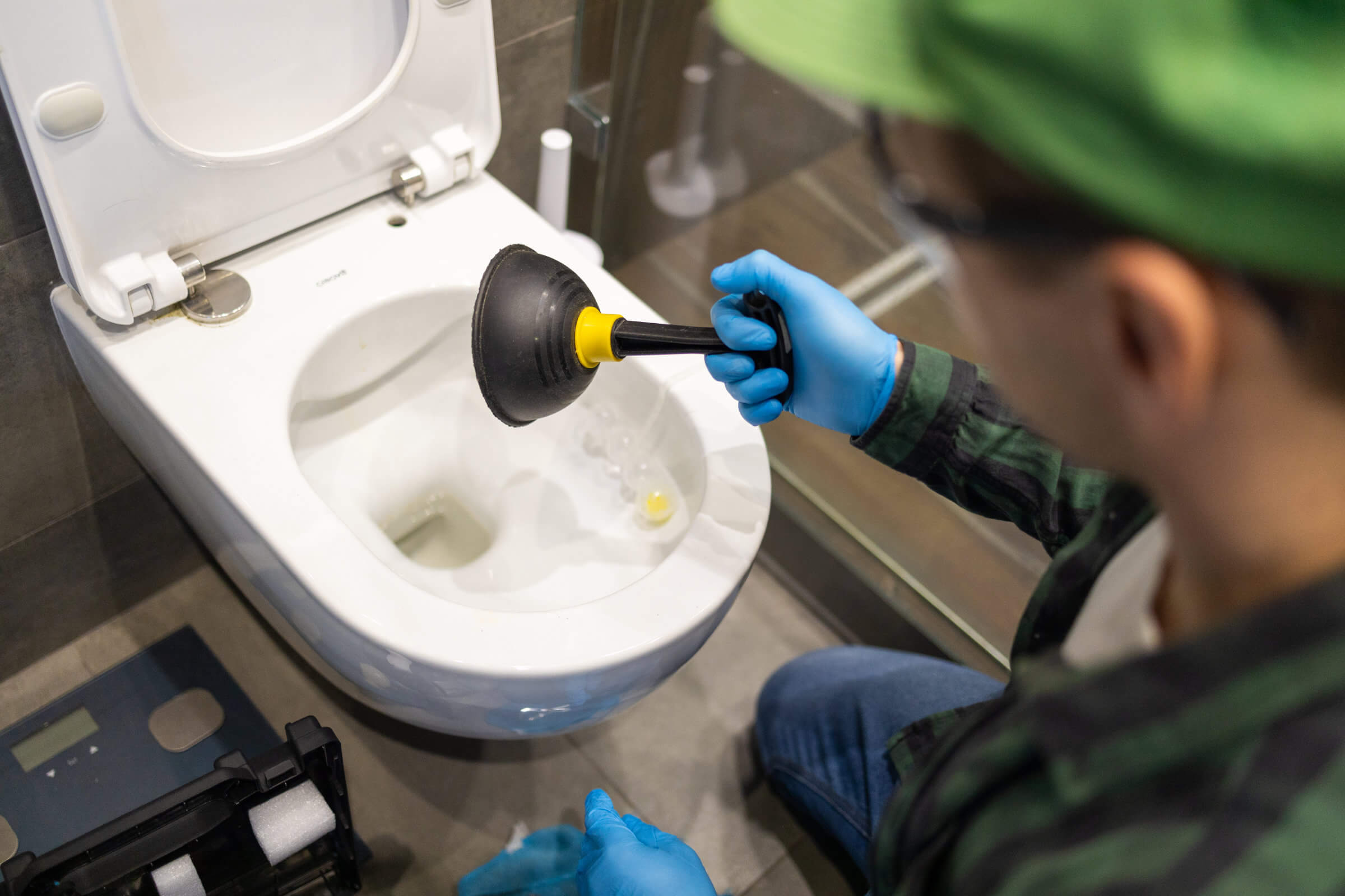 Description: Person in green hat and blue gloves using a plunger to unclog a blocked drain.